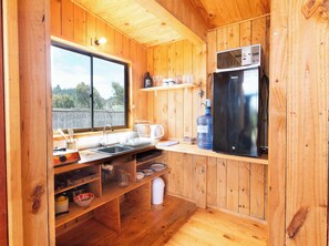 Une cuisine compacte aux murs en bois, dotĂ©e dâun rĂ©frigĂ©rateur en acier inoxydable, dâun Ă©vier et dâune fenĂȘtre donnant sur un paysage naturel.