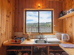 Une cuisine rustique amĂ©nagĂ©e dans une cabane en bois, avec une grande fenĂȘtre, des Ă©tagĂšres en bois, un Ă©vier et les appareils de cuisine de base.