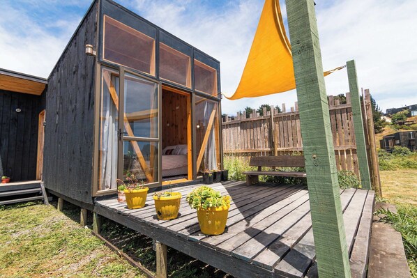 A wooden cabin with large windows, a deck with potted plants, and a wooden fence in the background.