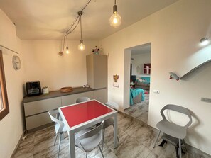 A dining area with a red table, grey chairs, a kitchen counter, and a living area with a sofa and a TV. - H 51 Family House: Your Ideal Vacation Home in Central Budoni (Budoni)