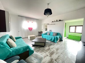 Een zithoek met een bank, voetenbankje en een salontafel. Er is een raam met gordijnen, een kroonluchter en een groene kast.