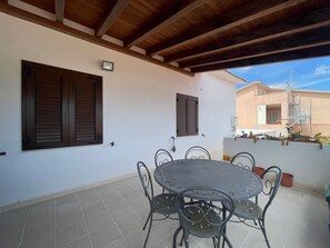 A covered patio with a round table and metal chairs, wooden beams, and a view of a building with a balcony. - H 51 Family House: Your Ideal Vacation Home in Central Budoni (Budoni)