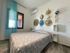 Un dormitorio con cama, ventana con cortinas, equipo de aire acondicionado y un perchero con sombreros.