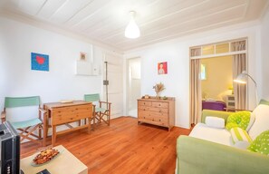 A room with a wooden floor, a green sofa, a wooden table, a dresser, and a bed with a purple bedspread.