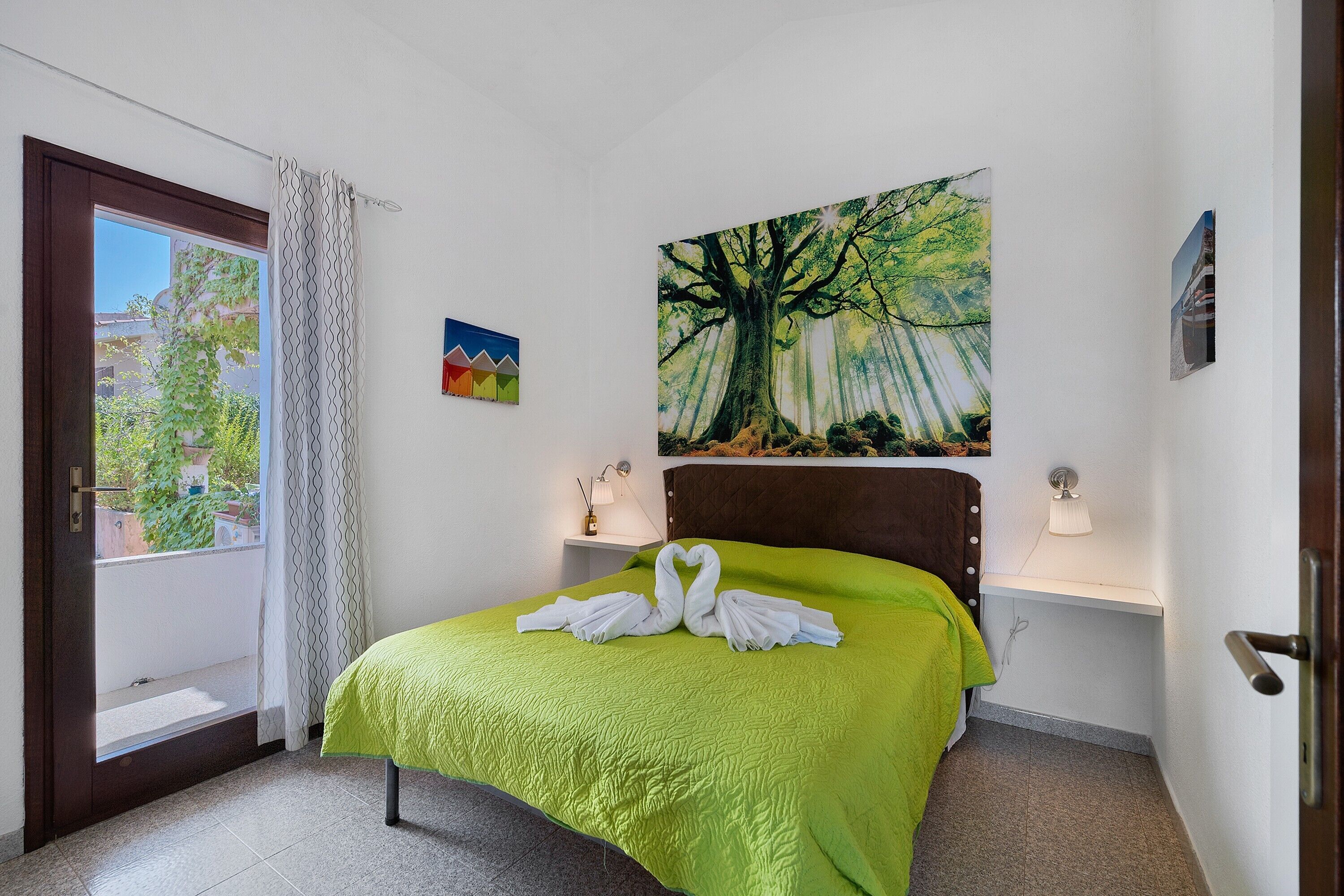 Une chambre d’hôtel avec un lit, un tableau représentant un arbre, une fenêtre avec des rideaux et une vue sur la verdure à l’extérieur.