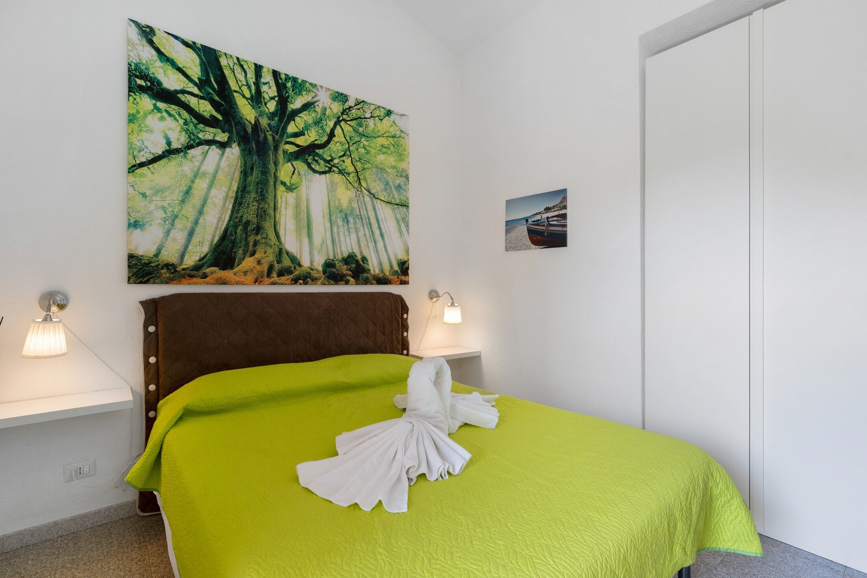 Habitación de hotel con cama, un cuadro de un árbol, una pequeña foto en la pared y una mesita de noche con lámpara.