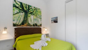Une chambre d’hôtel avec un lit, un tableau représentant un arbre, un petit cadre accroché au mur et une table de chevet avec une lampe.