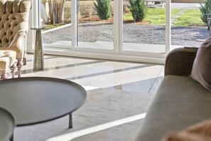 Ein modernes Wohnzimmer mit einer Couch, einem runden Tisch und einer großen Glasschiebetür zu einem Außenbereich mit Kiesweg und Grünflächen.