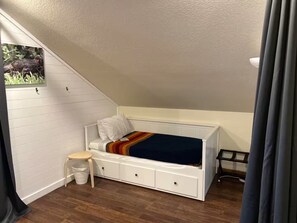 Ein kleines Schlafzimmer im Loft-Stil mit schrägen Wänden, einem Einzelbett, einem kleinen Hocker und einem Mülleimer.