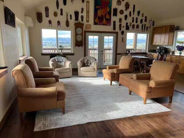 Ein geräumiges Wohnzimmer mit verschiedenen Sitzgelegenheiten, einem großen Fenster mit Blick und einem durchgehenden Türrahmen erkennbaren Küchenbereich.