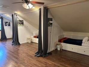Ein Schlafzimmer mit geneigter Decke, einem Deckenventilator, einem Bett mit farbenfroher Decke, einem kleinen Hocker und einer Stehlampe.