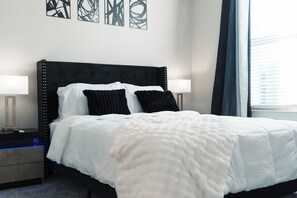 En pænt redt seng med hvidt og sort sengetøj, to sengeborde med lamper og abstrakt vægkunst.