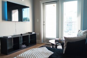 Una sala moderna con un televisor de pantalla plana, un mueble de entretenimiento negro, una mesa de centro redonda y un ventanal con persianas.