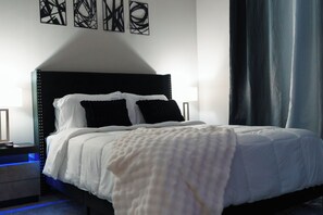 A neatly made bed with white bedding and black pillows, a bedside table with a lamp, and abstract wall art. - Modern Luxury ATL Apartment• World Cup 2026 (Atlanta)