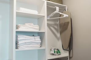 Een witte kast met planken waarop gevouwen handdoeken en een strijkplank met strijkijzer staan.