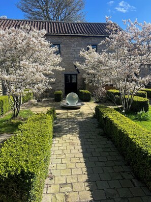 En bygning i sten med tegltag, en belagt sti og blomstrende træer.
