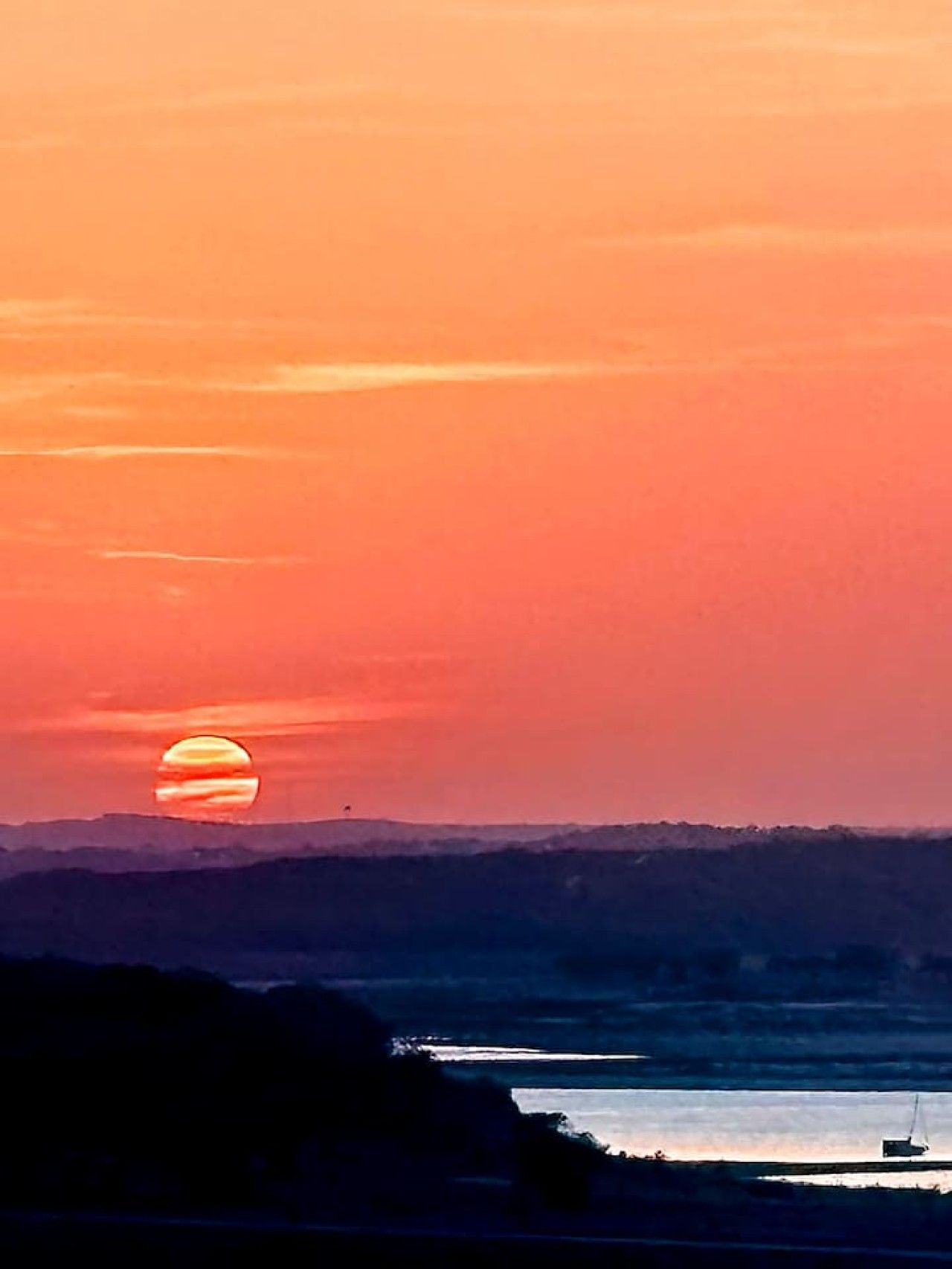 Een zonsondergang boven het water, met de silhouetten van een heuvel en een boot.
