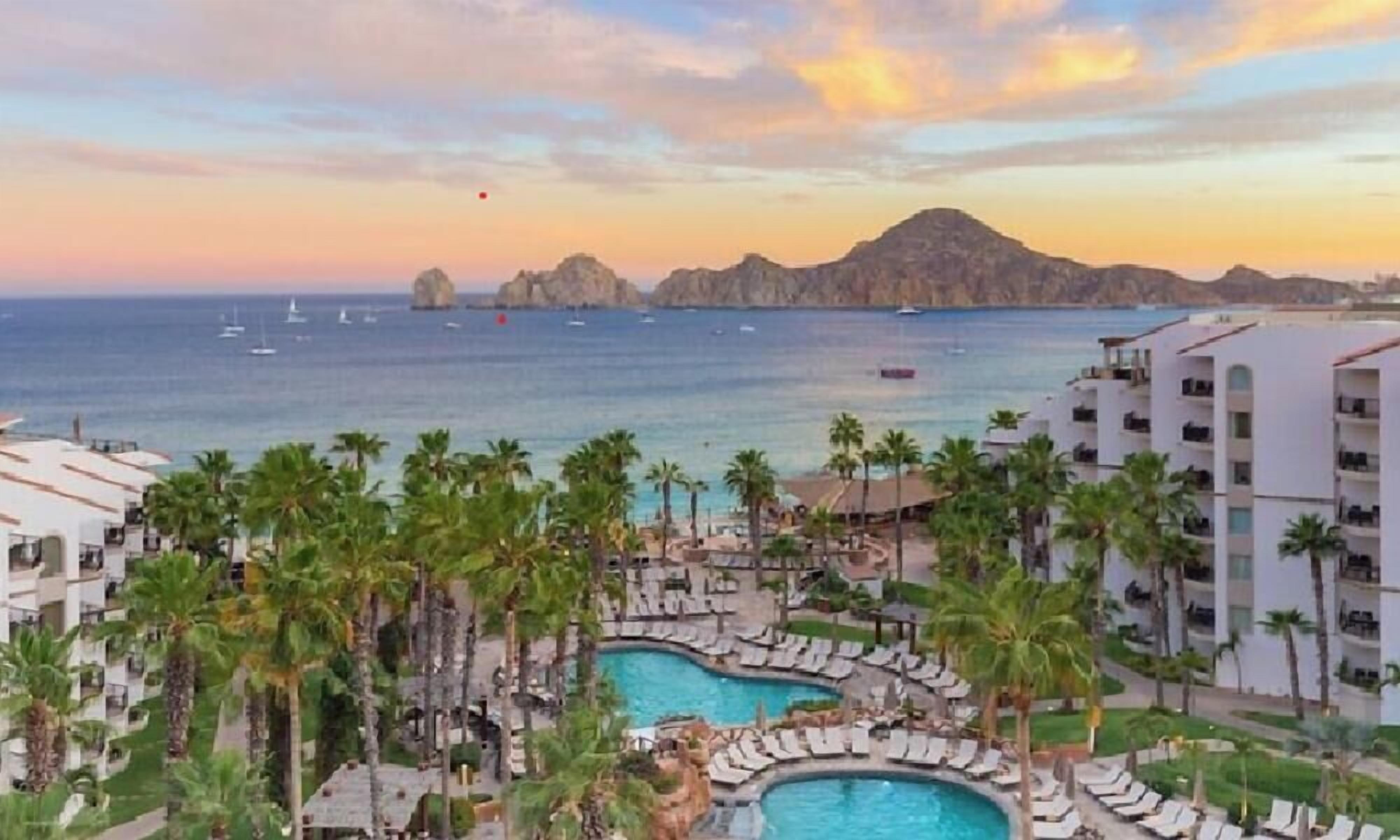 Een resort met een zwembad, palmbomen en uitzicht op zee bij zonsondergang.