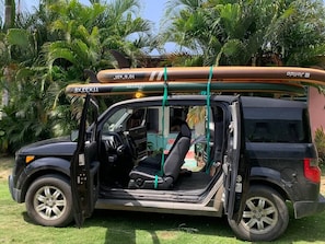 A vehicle with a surfboard strapped to the roof, parked in a grassy area with palm trees in the background.