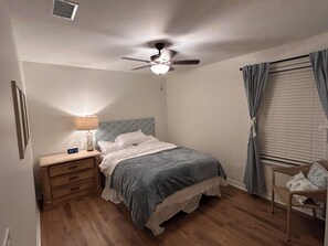A bedroom with a bed, nightstand, lamp, ceiling fan, window with curtains, and a chair. - Home with private pool near historic area and the beach in Fernandina Beach (Fernandina Beach)