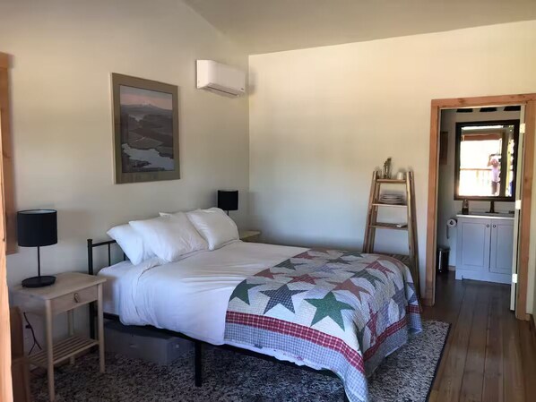 Ein Schlafzimmer mit Bett, Nachttisch, Lampe, einer Leiter, einem Gemälde und einer kleinen Küchenzeile.