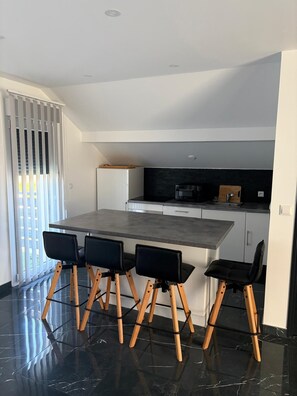 Een moderne keuken met een donker aanrecht, vier zwarte stoelen met houten poten en een witte koelkast.