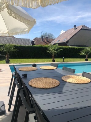 Zona de piscina al aire libre con una mesa y sillas, rodeada de vegetación y una casa al fondo.