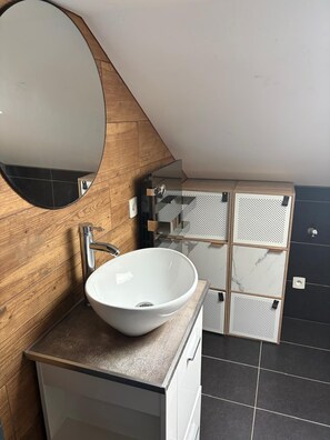 Un baño moderno con un espejo ovalado grande, un lavabo tipo recipiente blanco y una pared de madera.