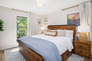 Ein Schlafzimmer mit einem hölzernen Bett, weißer Bettwäsche, einem Nachttisch mit Lampe und einem Fenster mit Vorhängen.