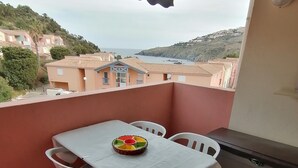 A balcony with a table and chairs, a wicker basket, and a view of buildings and hills. - Holiday Home in 'Port Aloès' Resort with Sea View, Shared Pool and Air Conditioning (Cerbère)