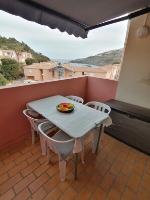 Un balcón con mesa y sillas, una cesta de mimbre y vistas a edificios y colinas.