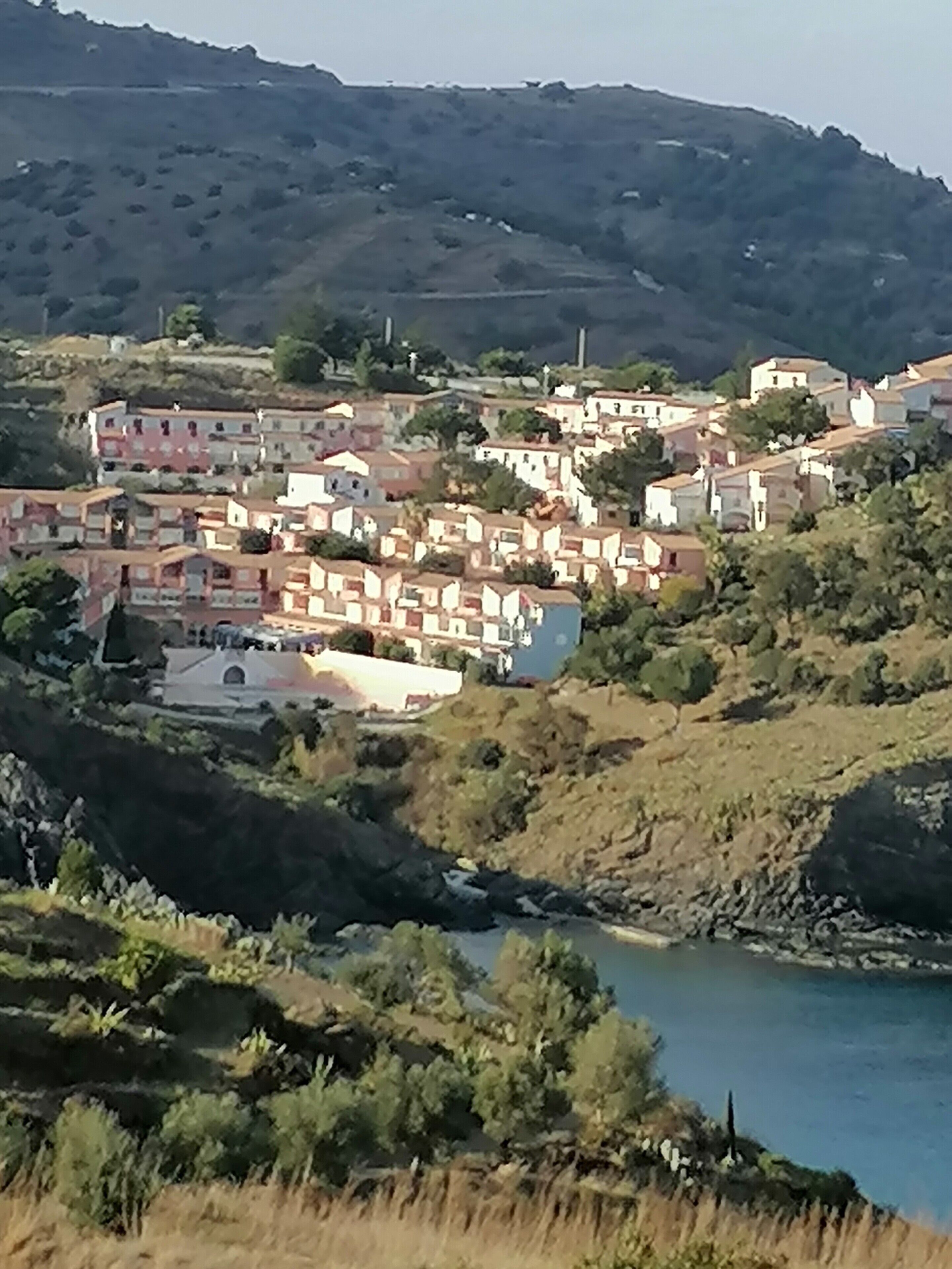 A hillside town with buildings clustered together, a river in the foreground, and a hilly backdrop.