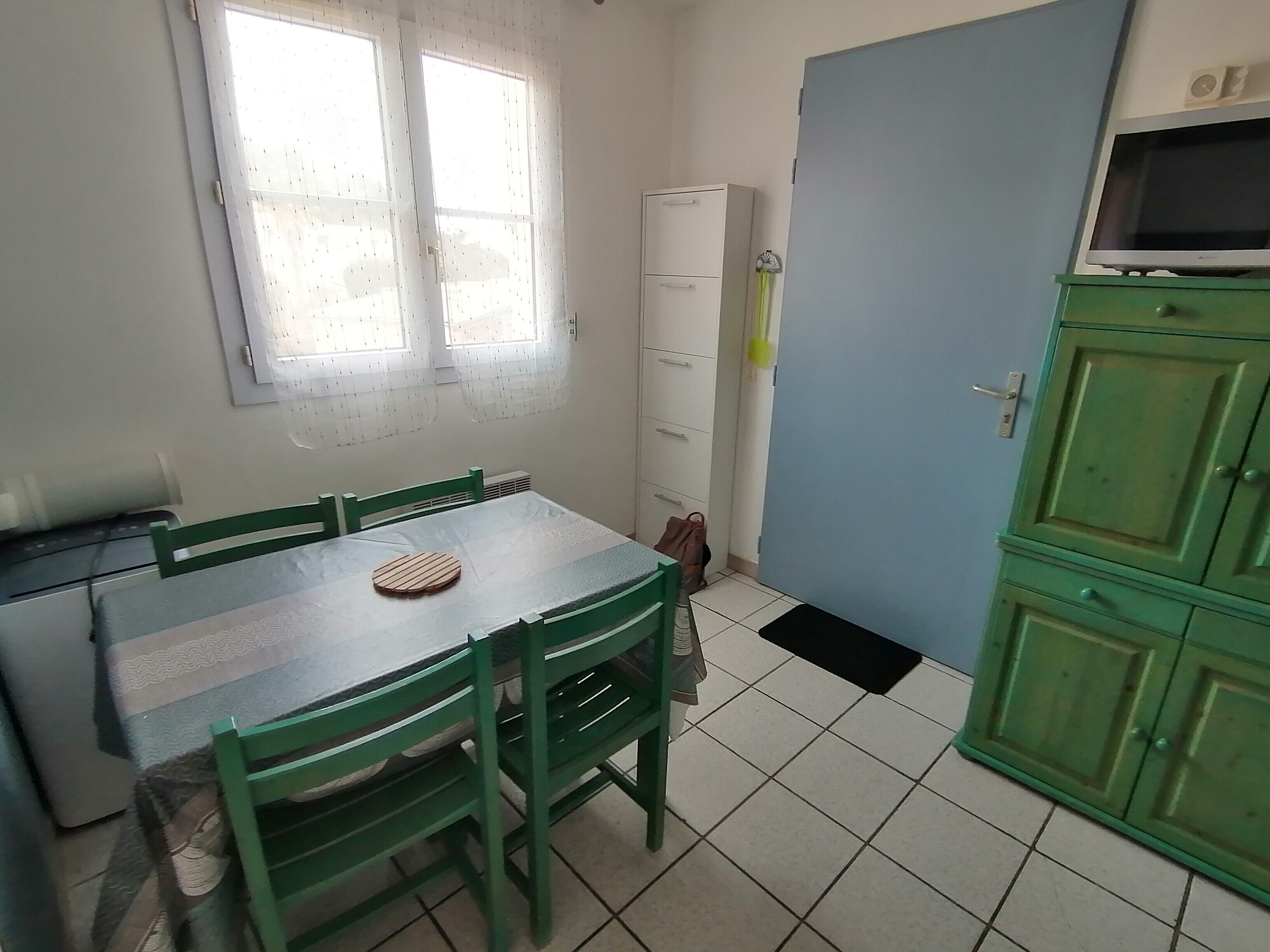 Ein Zimmer mit Esstisch, Stühlen, einem Fenster, einem weißen Schrank und einem grünen Schrank mit Fernseher.