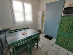 Una habitación con una mesa de comedor, sillas, un ventanal, un armario blanco y un mueble verde con un televisor.