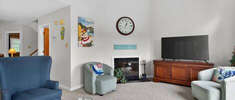 Una sala de estar con un sillón azul, un sillón gris, un televisor de pantalla plana, una chimenea y un reloj en la pared.
