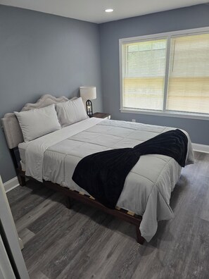 Quarto com cama, mesa de cabeceira, luminária e janela com persianas.