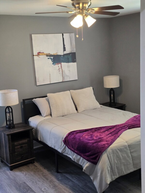 Um quarto com uma cama, duas mesas de cabeceira, um ventilador de teto, um quadro abstrato na parede e um cobertor roxo.