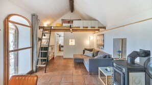 Un espacio habitable tipo loft con una cama en altillo, un sofá, una mesa de madera, una estufa y una escalera.