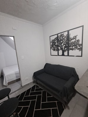 Een kleine, eenvoudig ingerichte kamer met een bank, een bed, een stoel en een zwart-wit tapijt.