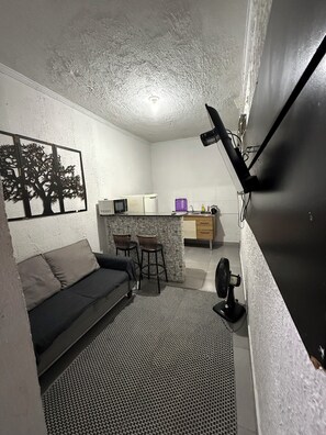 Un espacio habitable compacto con sofá, una pequeña zona de comedor, kitchenette y televisión montada en la pared.
