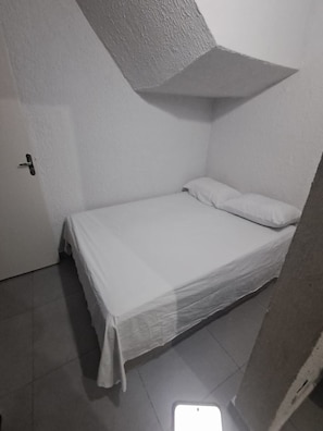 Een kleine kamer met een enkel bed, een effen witte muur, een deur en een betegelde vloer.
