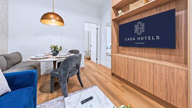 A modern room with a dining table set, a blue sofa, a grey armchair, a wooden cabinet, and a mounted TV displaying 'Casa Hotels Toronto'. - The Gerrard Brand New CN Tower Home Base (Toronto)