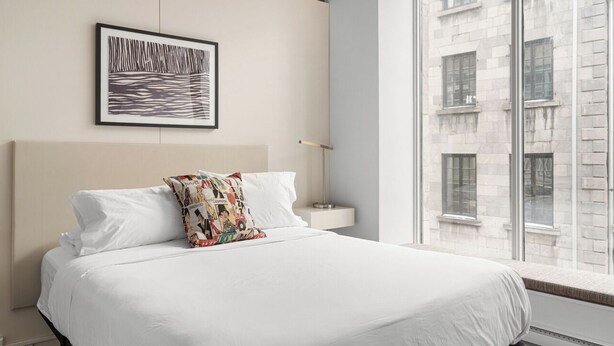 Ein Hotelzimmer mit einem großen Bett, einem gerahmten Bild an der Wand, einem Nachttisch und Blick auf ein Gebäude durch das Fenster.