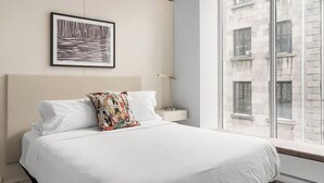 Ein Hotelzimmer mit einem großen Bett, einem gerahmten Bild an der Wand, einem Nachttisch und Blick auf ein Gebäude durch das Fenster.