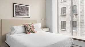 A hotel room with a large bed, a framed picture on the wall, a bedside table, and a view of a building through the window.