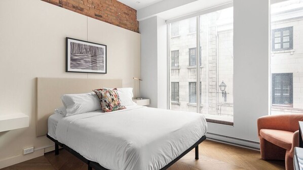 A bedroom with a bed, a window, a chair, and a brick wall.