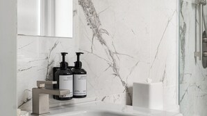 Ein Badezimmer mit Marmorwand, einem weißen Waschbecken, zwei schwarzen Flaschen mit Körperlotion und einer Duschkabine aus Glas.