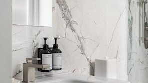 Ein Badezimmer mit Marmorwand, einem weißen Waschbecken, zwei schwarzen Flaschen mit Körperlotion und einer Duschkabine aus Glas.