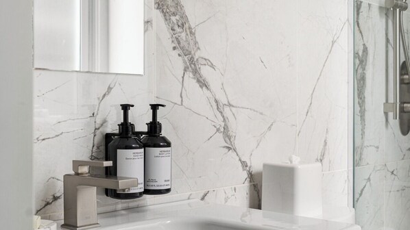 A bathroom with a marble wall, a white sink, two black bottles of body lotion, and a glass shower enclosure. - Bowery Lofts Hotel Vieux Montreal (Montréal)