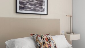 Ein Hotelzimmer mit Bett, einem Kissen mit farbigem Muster, einem gerahmten Kunstwerk über dem Bett und einem Nachttisch mit Lampe.